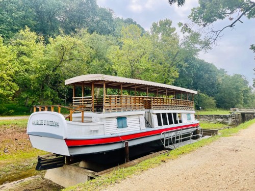 A white houseboat with a red stripe is docked near a tree-lined path and a lock in a serene outdoor setting.