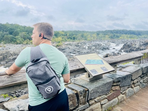 A person with a backpack stands by a railing, looking at a river and rocky landscape under a cloudy sky.