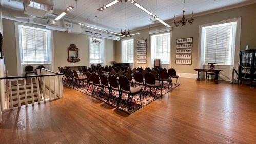 A spacious room with wooden floors, arranged chairs, and historical decor, featuring large windows and a rug.