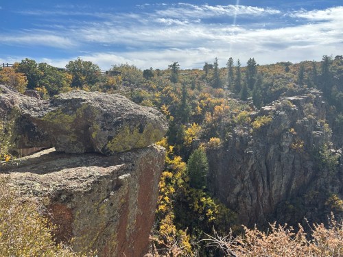A rocky cliff edge overlooks a vibrant forest with autumn foliage under a blue sky with wispy clouds.