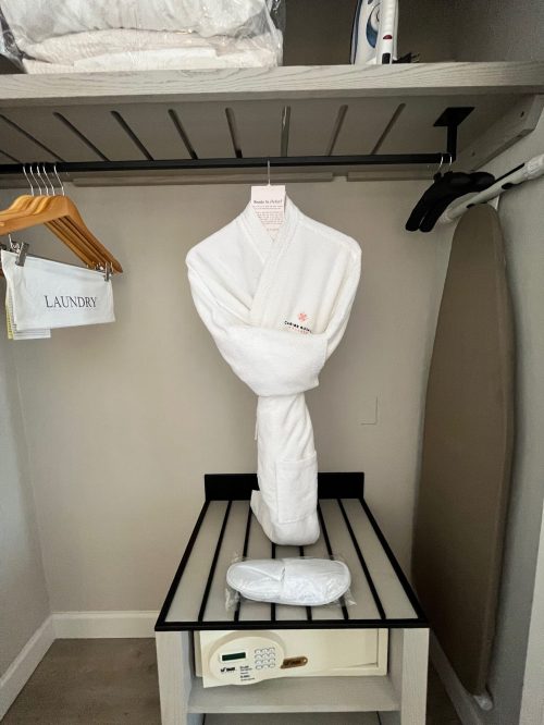 A white bathrobe hangs in a closet next to a laundry bag and an iron, with a safe on a black and white table below.