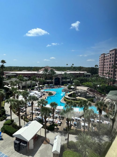 A sunny view of a resort pool surrounded by palm trees and lounge chairs, with a hotel in the background.