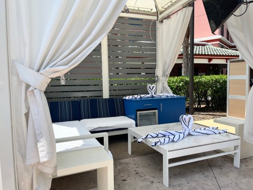A cozy outdoor cabana with white curtains, a blue cabinet, and towel swans on the seating area.