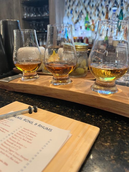 Three glasses of rum in varying shades of amber on a wooden flight board, with a menu in the foreground.