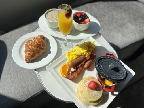 A breakfast spread featuring pancakes, eggs, bacon, sausage, a croissant, fruit, oatmeal, and a mimosa.