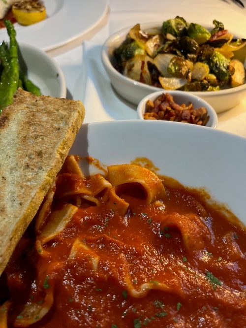 A plate of pasta in tomato sauce with toast, accompanied by Brussels sprouts and a small bowl of bacon bits.