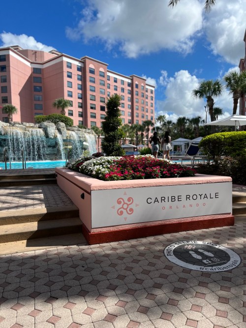 Sign for Caribe Royale Orlando in front of a pool area with flowers and a waterfall, surrounded by palm trees.