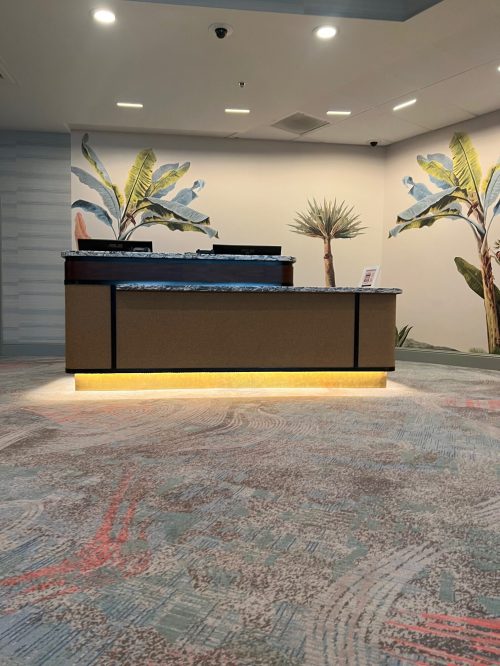 A modern reception desk with a granite top, surrounded by tropical plant murals and patterned carpet.