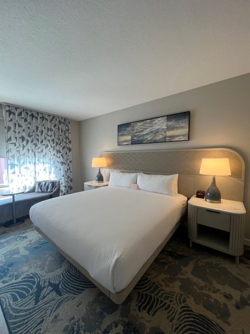 A modern hotel room featuring a large bed, bedside tables, lamps, and a window with patterned curtains.