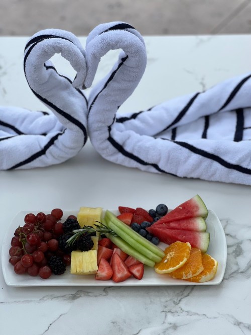 Two towel swans form a heart shape behind a colorful fruit platter featuring grapes, berries, pineapple, and watermelon.