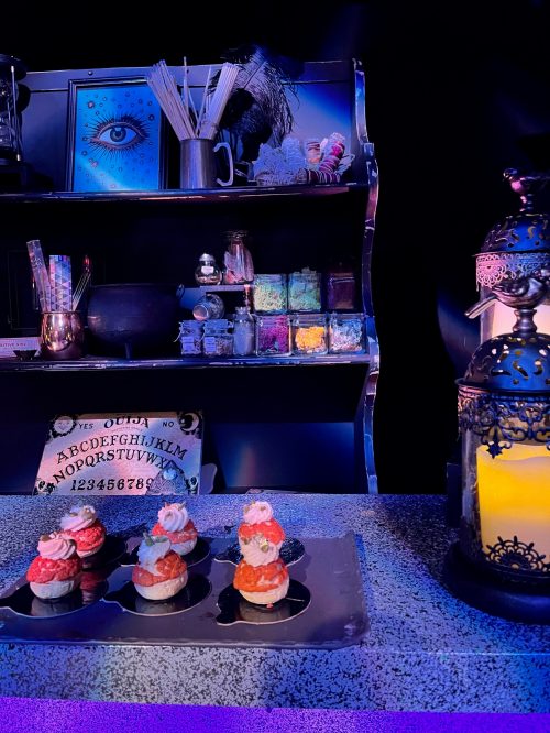 A display of colorful cupcakes on a table, with a decorative shelf and lantern in a dimly lit setting.