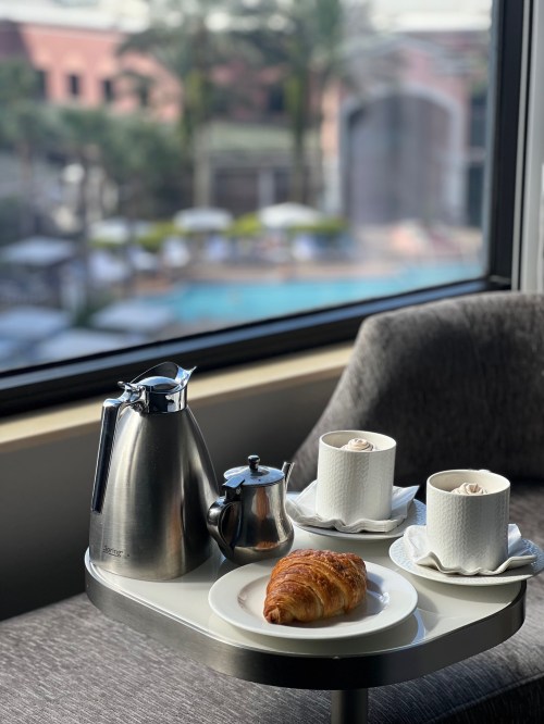 A cozy breakfast setup with a silver kettle, two cups, a croissant, and a view of a pool outside.