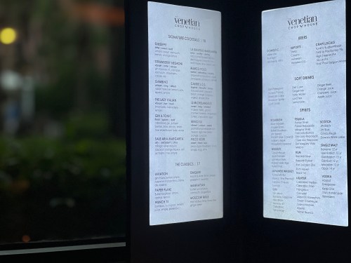 Menu boards displaying cocktails, beers, soft drinks, and spirits at the Venetian Copy House, illuminated in soft light.