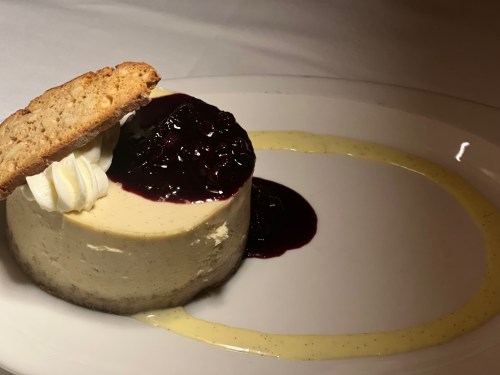 A slice of cheesecake topped with berry sauce, whipped cream, and a cookie, served on a white plate with a sauce drizzle.