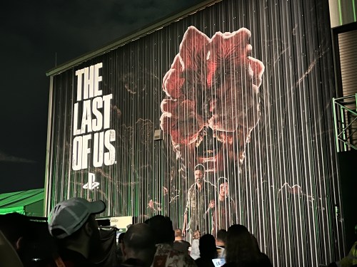 A large mural of "The Last of Us" featuring characters Joel and Ellie, with a crowd in front at night.