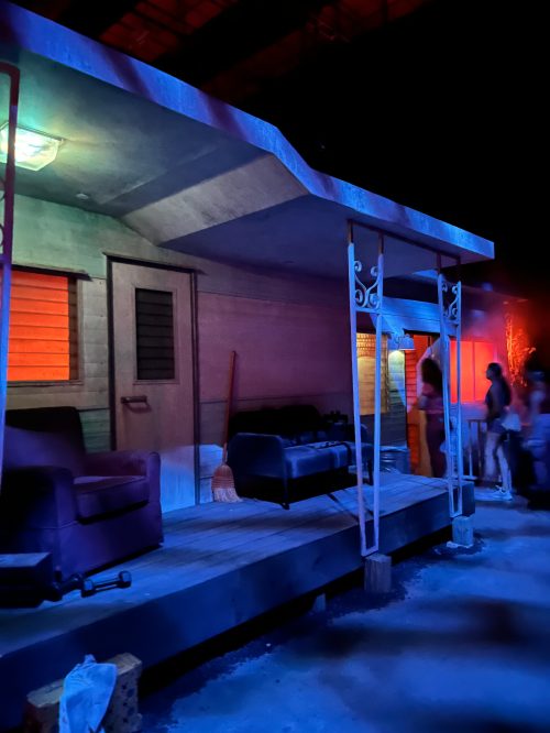 A dimly lit set featuring a porch with two sofas, colorful lighting, and a broom on the ground.
