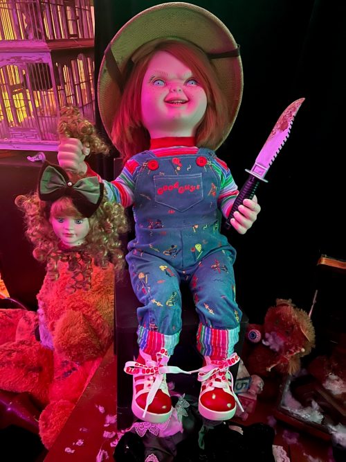 A doll resembling a horror character holds a knife and a severed head, surrounded by colorful, chaotic decor.