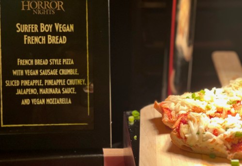 A close-up of Surfer Boy Vegan French Bread pizza with toppings, next to a sign detailing its ingredients.
