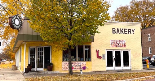 A yellow bakery building with large windows, an open sign, and autumn leaves surrounding it. The word "BAKERY" is prominently displayed.