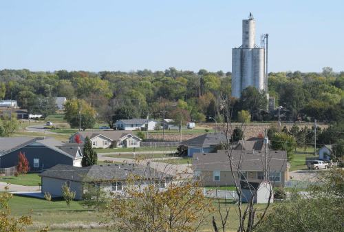 A rural landscape featuring houses, trees, and a tall grain silo in the background under a clear blue sky.