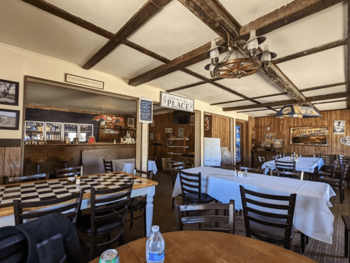Cozy restaurant interior with wooden beams, checkered table, and empty tables set for dining. Bar area in the background.