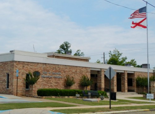 Exterior of a brick municipal building with a flagpole displaying the American and Alabama flags. Green landscaping in front.