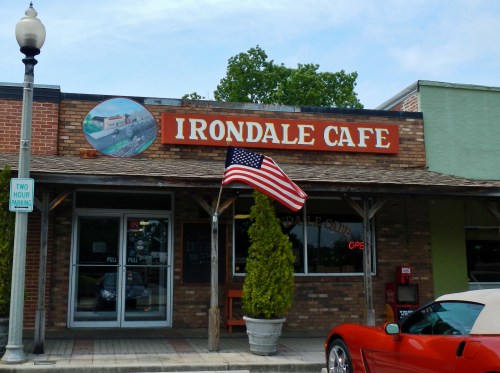 Exterior of Irondale Cafe with an American flag, a red sports car parked nearby, and a sign indicating open hours.