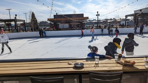 A busy outdoor ice skating rink with people skating and a table in the foreground with drinks and snacks.