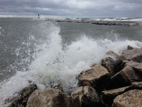 Waves crash against rocky shore with a green lighthouse in the distance under a cloudy sky.