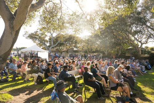 A large crowd sits on benches in a sunny outdoor setting, surrounded by trees and a festive atmosphere.
