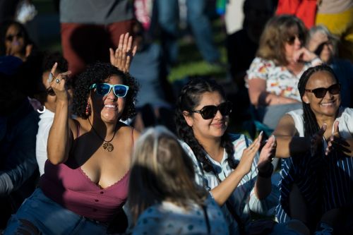 A diverse group of women enjoying an outdoor event, smiling, clapping, and wearing sunglasses.