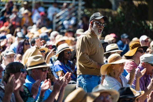 A diverse crowd of people wearing hats, some clapping and smiling, enjoying an outdoor event.