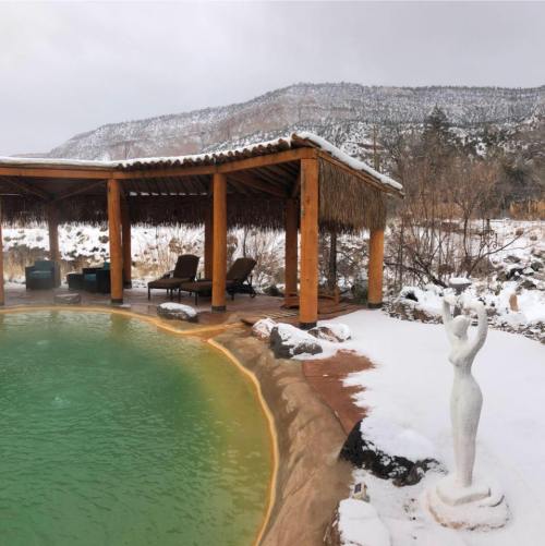 A serene outdoor spa with a green pool, surrounded by snow and mountains, featuring a statue and wooden shelter.