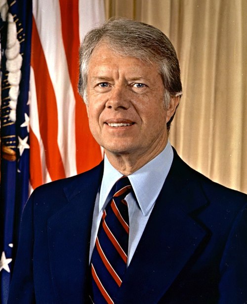 Portrait of a man in a suit with a blue shirt and striped tie, standing in front of an American flag backdrop.