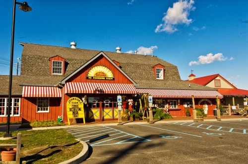 A red barn-style building with a welcoming sign, striped awnings, and a parking lot on a sunny day.