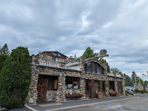 A rustic stone building with a sign reading "Lago Inn," surrounded by greenery and under a cloudy sky.
