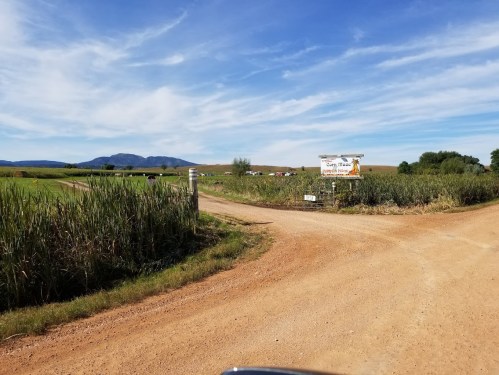 A dirt road splits at a fork, leading to fields and a sign for a farm, with mountains in the background under a blue sky.