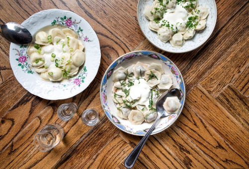 Three bowls of dumplings topped with sour cream and herbs, served on a wooden table with shot glasses nearby.