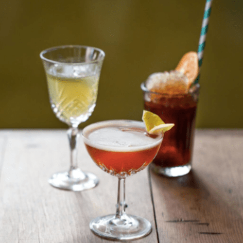 Three cocktails on a wooden table: a clear drink, a dark drink with a straw, and a reddish drink with a lemon slice.