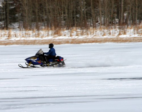 A person riding a snowmobile across a frozen lake, surrounded by trees and snow.
