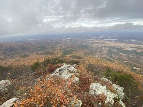 A panoramic view from a mountain peak, showcasing rolling hills and autumn foliage under a cloudy sky.