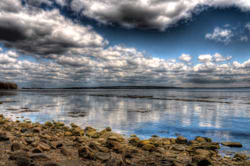 A serene lakeside view with rocky shore, calm water reflecting clouds, and a distant horizon under a blue sky.