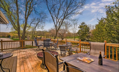 A sunny deck with tables and chairs, featuring a wine bottle and snacks, surrounded by trees and a clear blue sky.