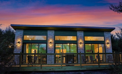 Modern cabin with large windows, wooden deck, and stone accents, set against a colorful sunset sky.
