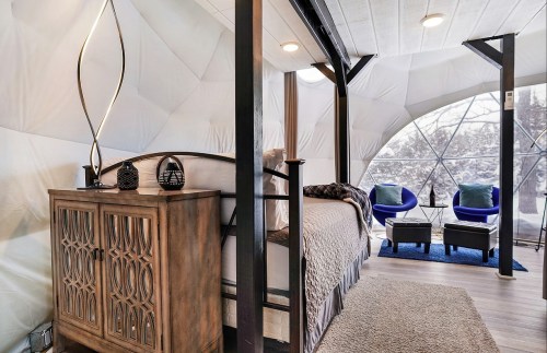 Cozy interior of a geodesic dome with a bed, wooden cabinet, and seating area, surrounded by large windows.