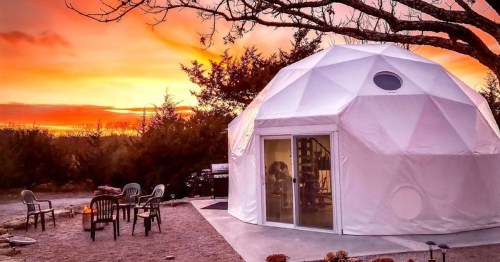 A geodesic dome home at sunset, surrounded by trees and outdoor seating. Warm colors fill the sky.