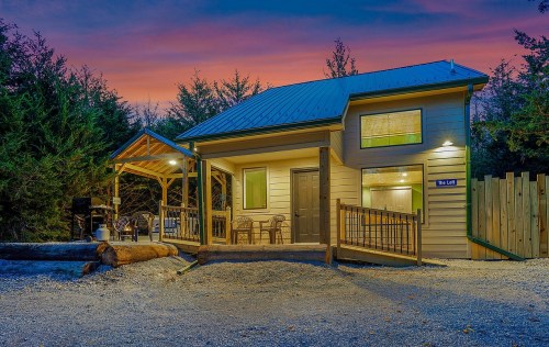 A cozy cabin with a porch, surrounded by trees, under a colorful sunset sky.