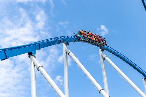 A roller coaster reaches the top of a steep hill against a blue sky, with riders enjoying the thrill.