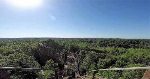 A panoramic view from the top of a wooden roller coaster, overlooking lush green trees and a clear blue sky.