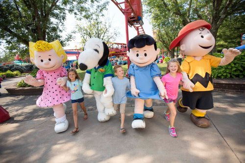 Three children hold hands with costumed characters from the Peanuts gang, smiling in a sunny outdoor setting.